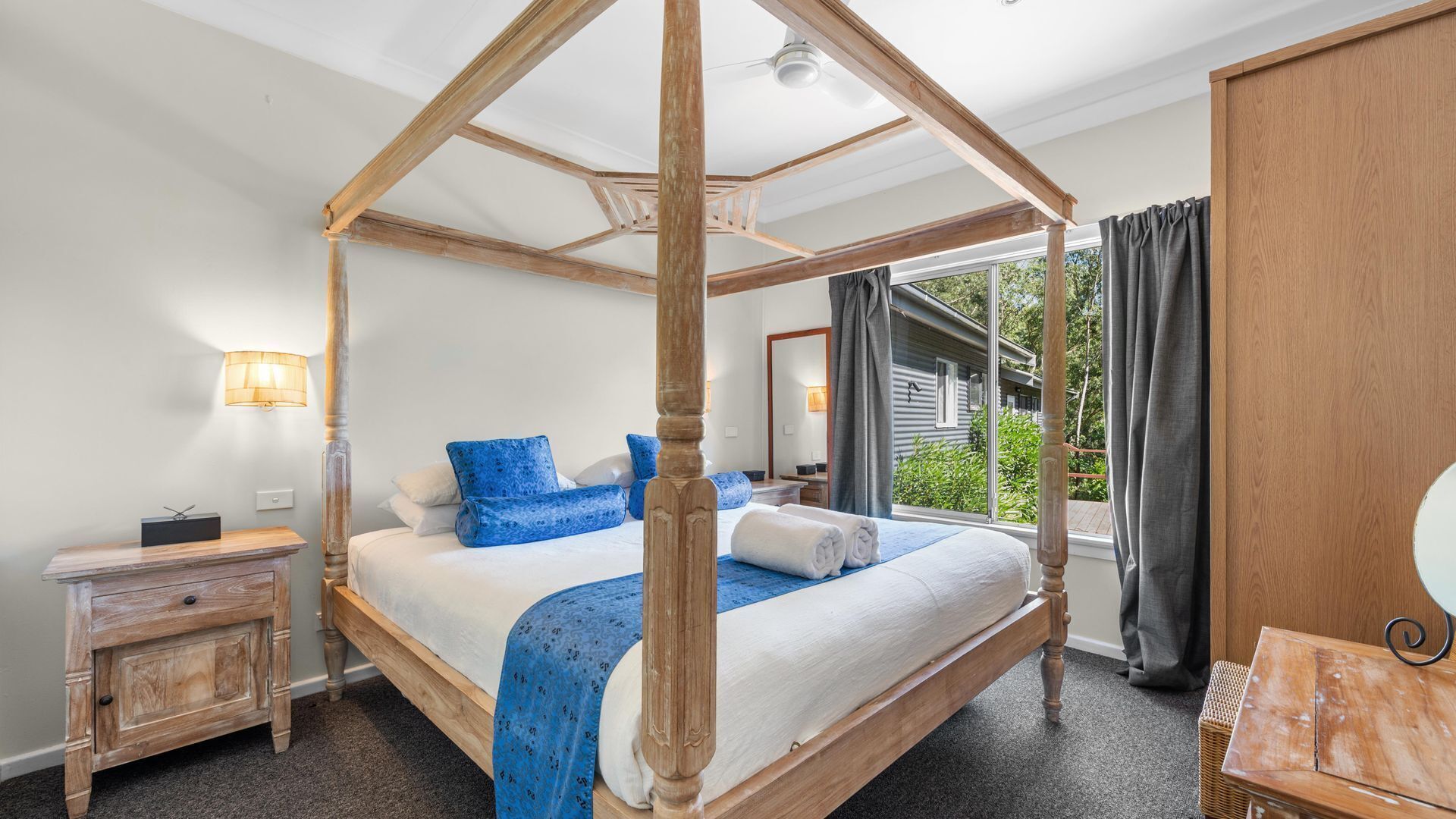 A four-poster canopy bed with a view of the outdoors, a bedside table, and a wooden wardrobe.