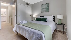 Une chambre à coucher avec un lit, des lampes de chevet, un plaid vert, une porte blanche et un tableau accroché au mur.