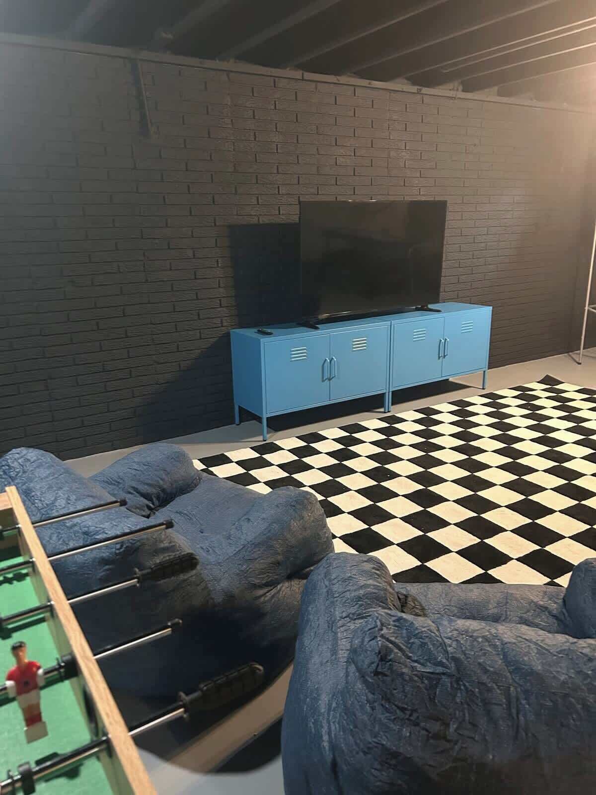 A room with a brick wall, a blue cabinet with a flat-screen TV, a checkered floor, and a foosball table.