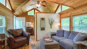 Ein Interieur einer Blockhütte mit einem braunen Ledersofa, einem grauen Sofa, einer Holzdeckenverkleidung mit Lüfter und einem Fenster mit Jalousien.