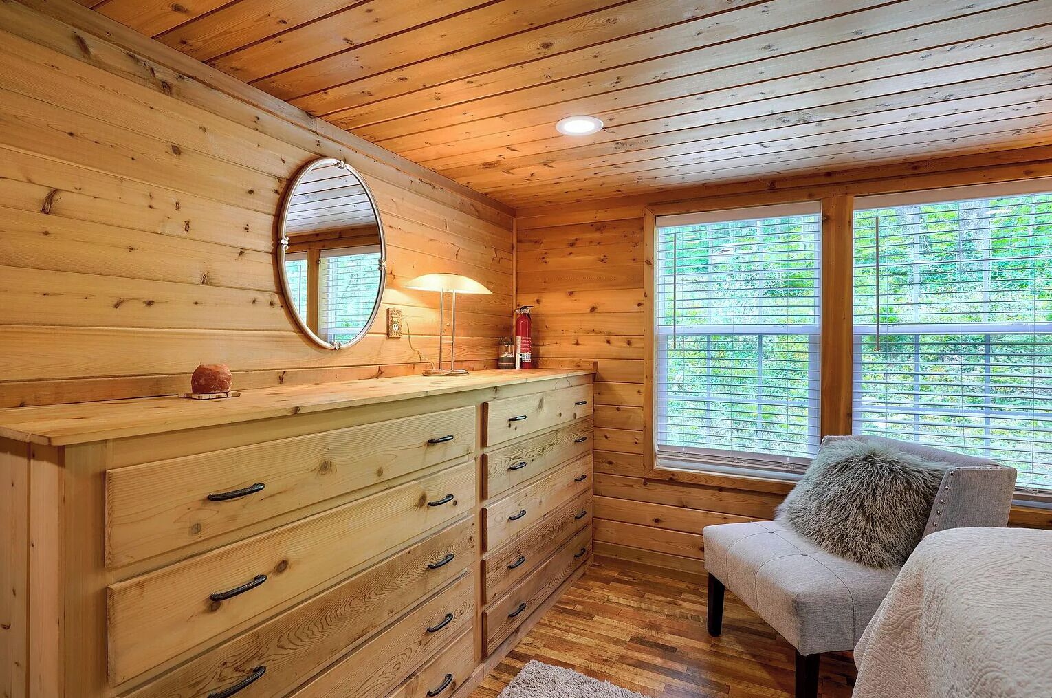 L’intérieur d’un chalet en bois, avec une grande commode en bois, un miroir, une fenêtre donnant sur la verdure et un coin salon confortable.