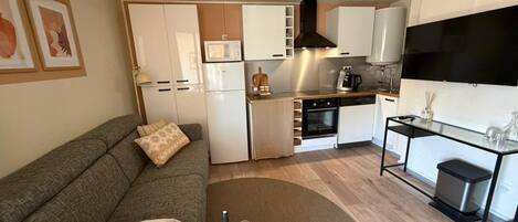 Un espacio habitable compacto con un sofá, una pequeña mesa de centro, una zona de cocina que incluye microondas, horno y un televisor.