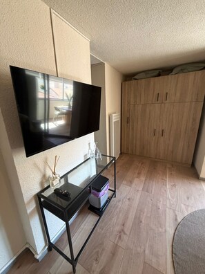Ein Wohnzimmer mit Flachbildfernseher an der Wand, einem Glastisch mit Fernbedienung, einer kleinen Vase und einer Aufbewahrungsbox.