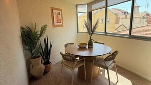 A room with a round wooden table, four wicker chairs, a potted plant, a framed picture on the wall, and a large window with a view of buildings. - Modern Beach Apartment Valras (Valras-Plage)