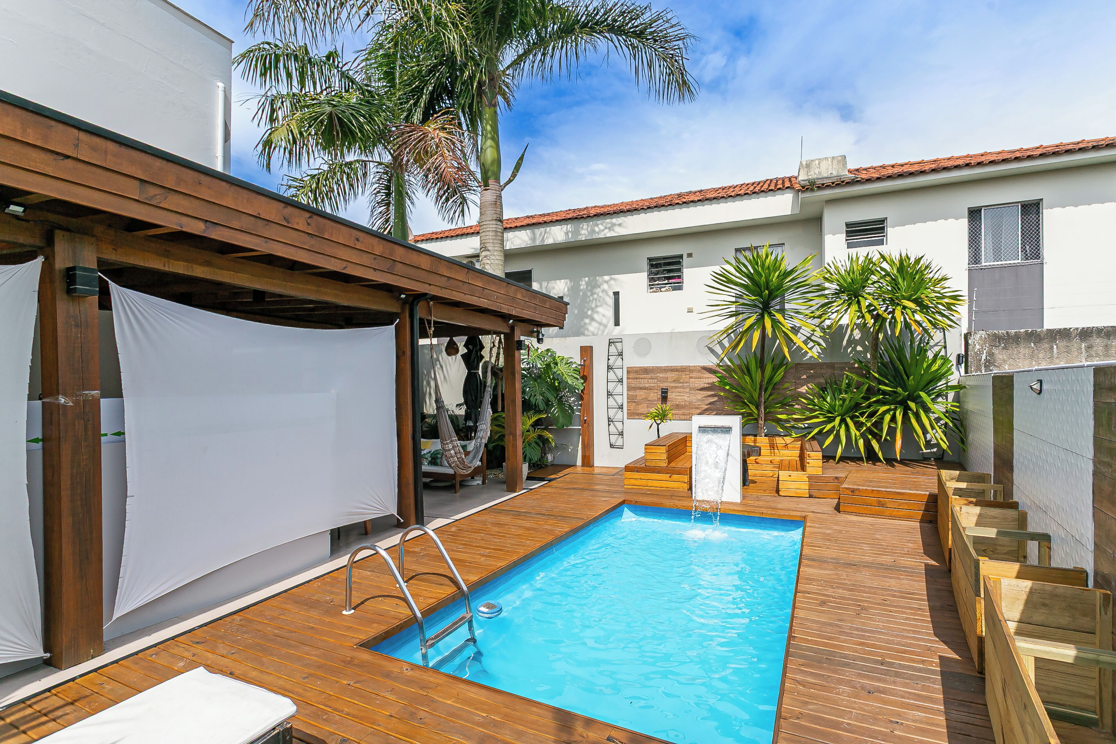 A wooden deck with a swimming pool, a covered seating area, and tropical plants.
