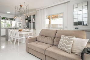 A modern living room with a beige sofa, a dining area with white chairs, and a kitchen in the background. - Elegante Casa com Piscina - Canasvieiras (Florianópolis)