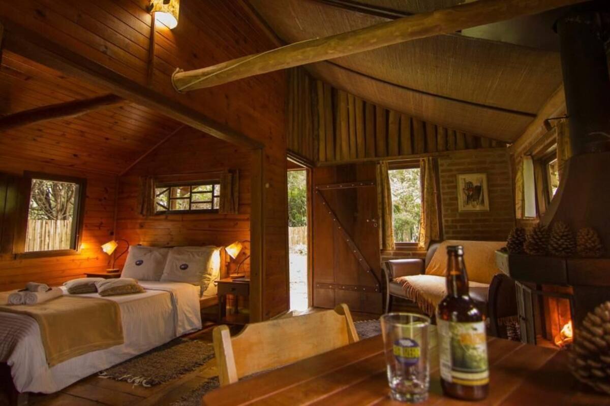 Acogedora habitación tipo cabaña de madera con cama, mesa con botellas, chimenea y un ventanal grande.