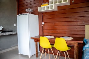 A compact kitchen with a white refrigerator, a wooden dining table with two yellow chairs, and a shelf above holding glasses and a small yellow object.