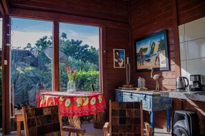 A wooden interior with a dining table, chairs, a painting, and a view of greenery outside.