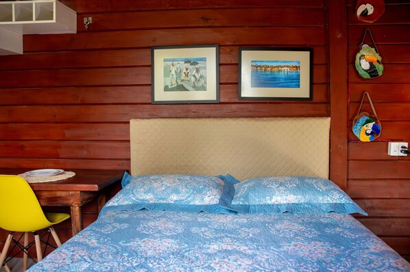 A room with a bed, a wooden wall, a table with a chair, and two framed pictures.