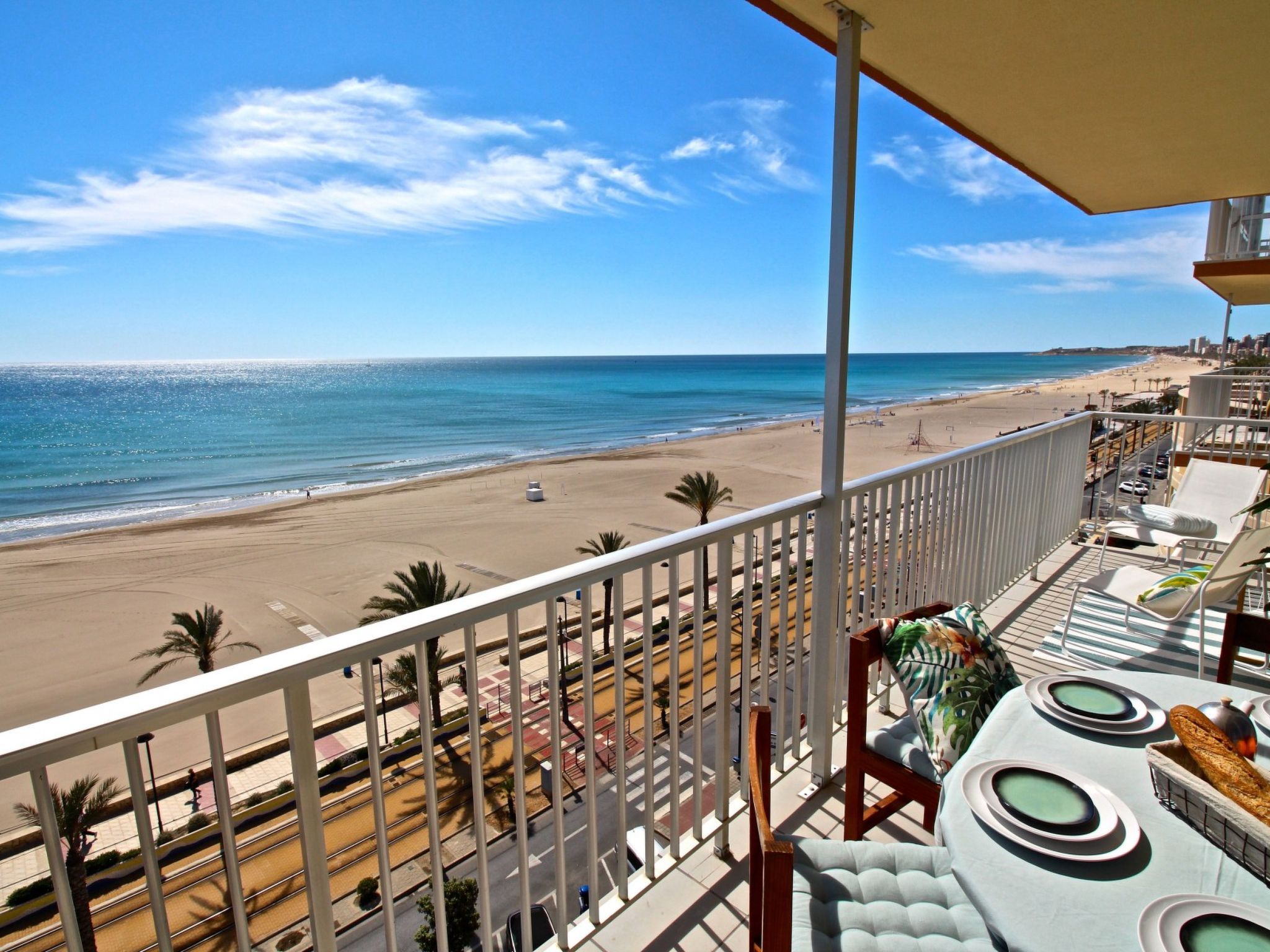 A balcony with a view of a beach and ocean, a dining area with a table and chairs, and a railing.