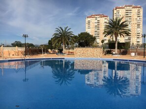 A swimming pool with a stone wall and palm trees, surrounded by a fence and buildings in the background. - ESTUDIO PLAYA MUCHAVISTA by Interhome (El Campello/Villajoyosa)