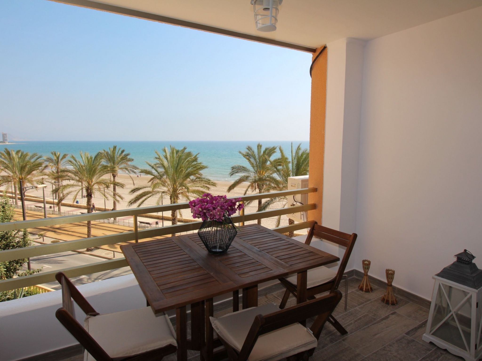 A balcony with a wooden table and chairs, a vase of flowers, and a view of the beach and ocean.