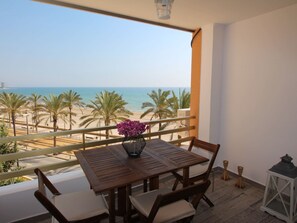 A balcony with a wooden table and chairs, a vase of flowers, and a view of the beach and ocean. - ESTUDIO PLAYA MUCHAVISTA by Interhome (El Campello/Villajoyosa)