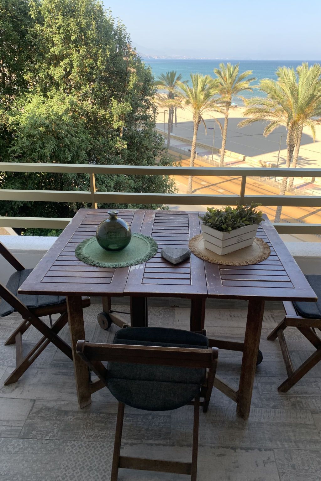 A wooden table with a green mat, a potted plant, and a stone on it, set on a balcony with a view of the sea and palm trees.