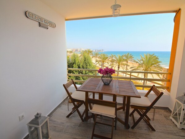 A balcony with a wooden table and chairs, a flower vase, and a view of the beach and ocean. - ESTUDIO PLAYA MUCHAVISTA by Interhome (El Campello/Villajoyosa)
