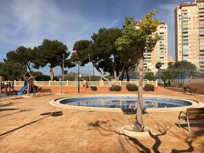 A public park with a circular pool, benches, trees, and a playground. - ESTUDIO PLAYA MUCHAVISTA by Interhome (El Campello/Villajoyosa)