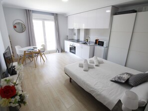 Ein modernes Hotelzimmer mit einem Bett, einer kleinen Küchenzeile, einem Essbereich mit Tisch und Stühlen sowie einem großen Fenster.