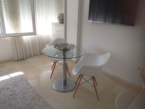 A modern dining area with a glass table and two white chairs, a white cabinet, and a window with blinds. - ESTUDIO PLAYA POSTIGUET by Interhome (Alicante)