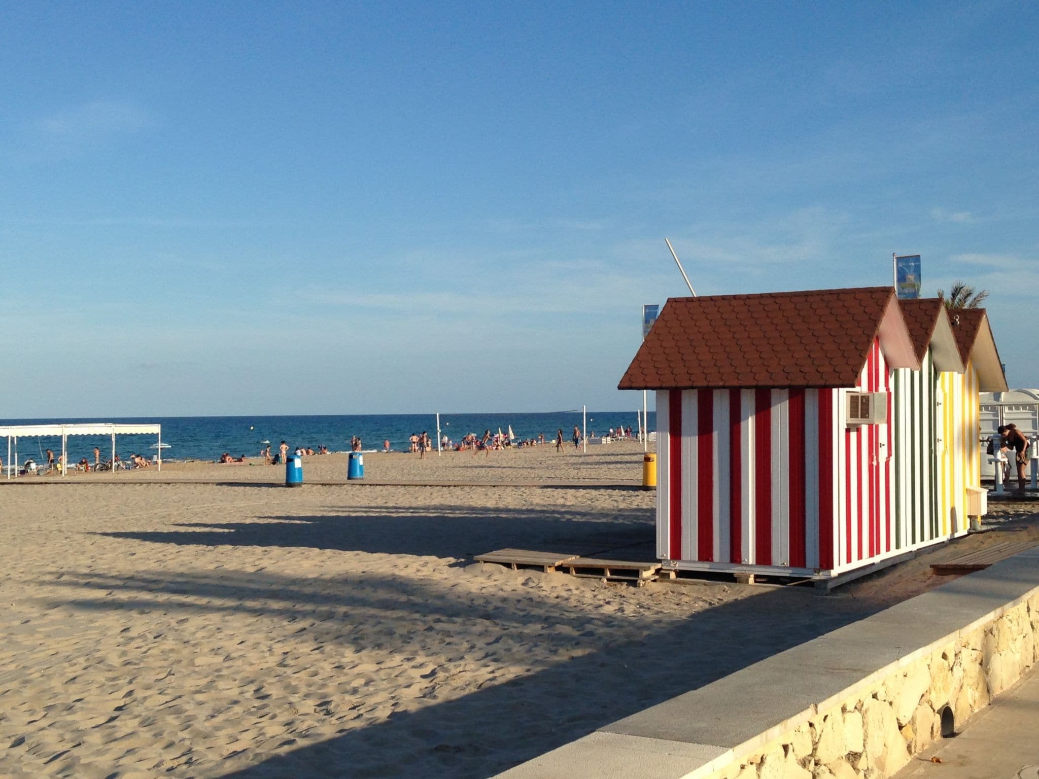 Ein Strand mit einer kleinen, gestreiften Strandhütte, einem überdachten Bereich und blauem Himmel.
