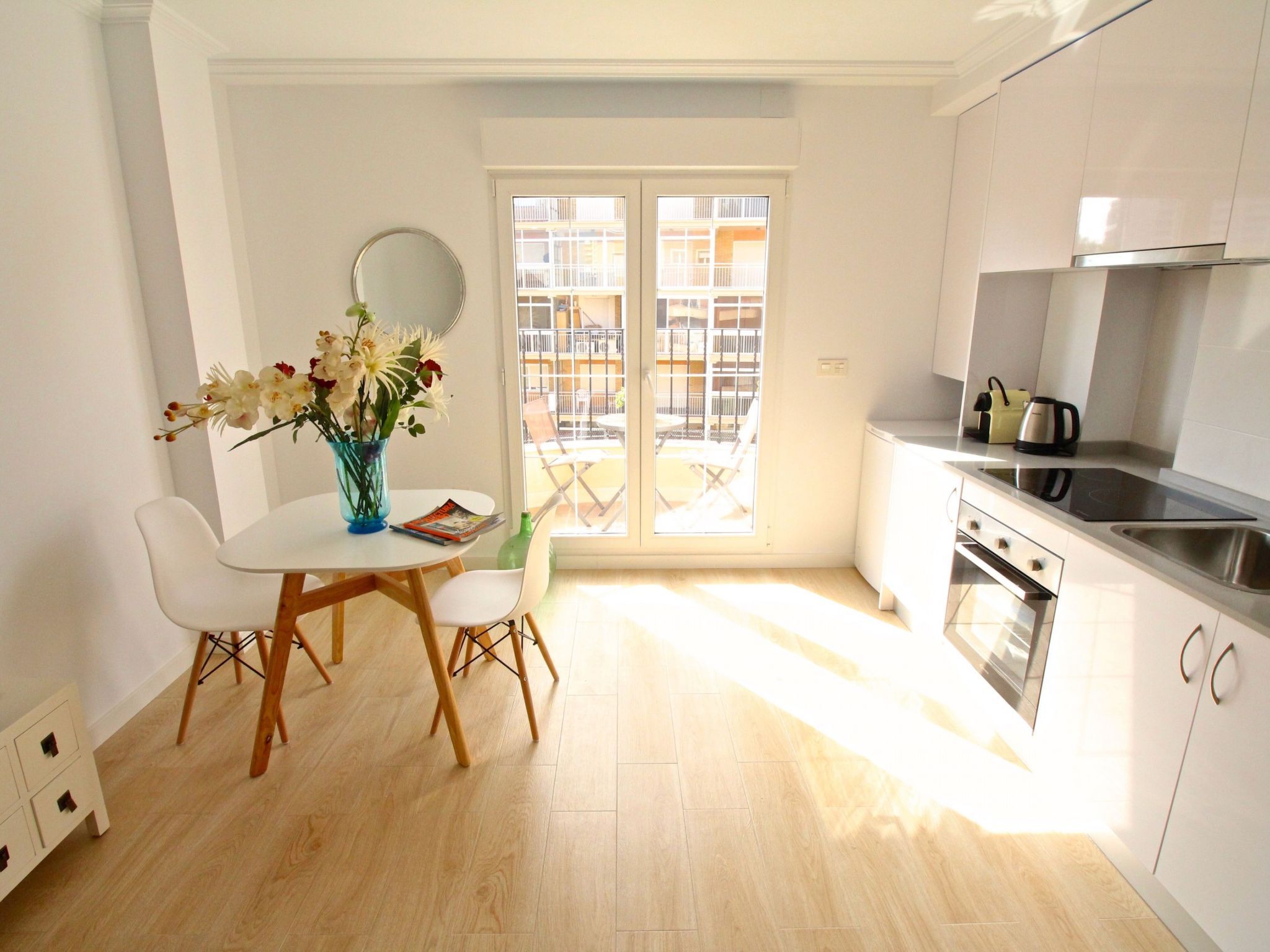 A modern kitchen with a dining area, a view through a glass door, and a vase with flowers on the table.