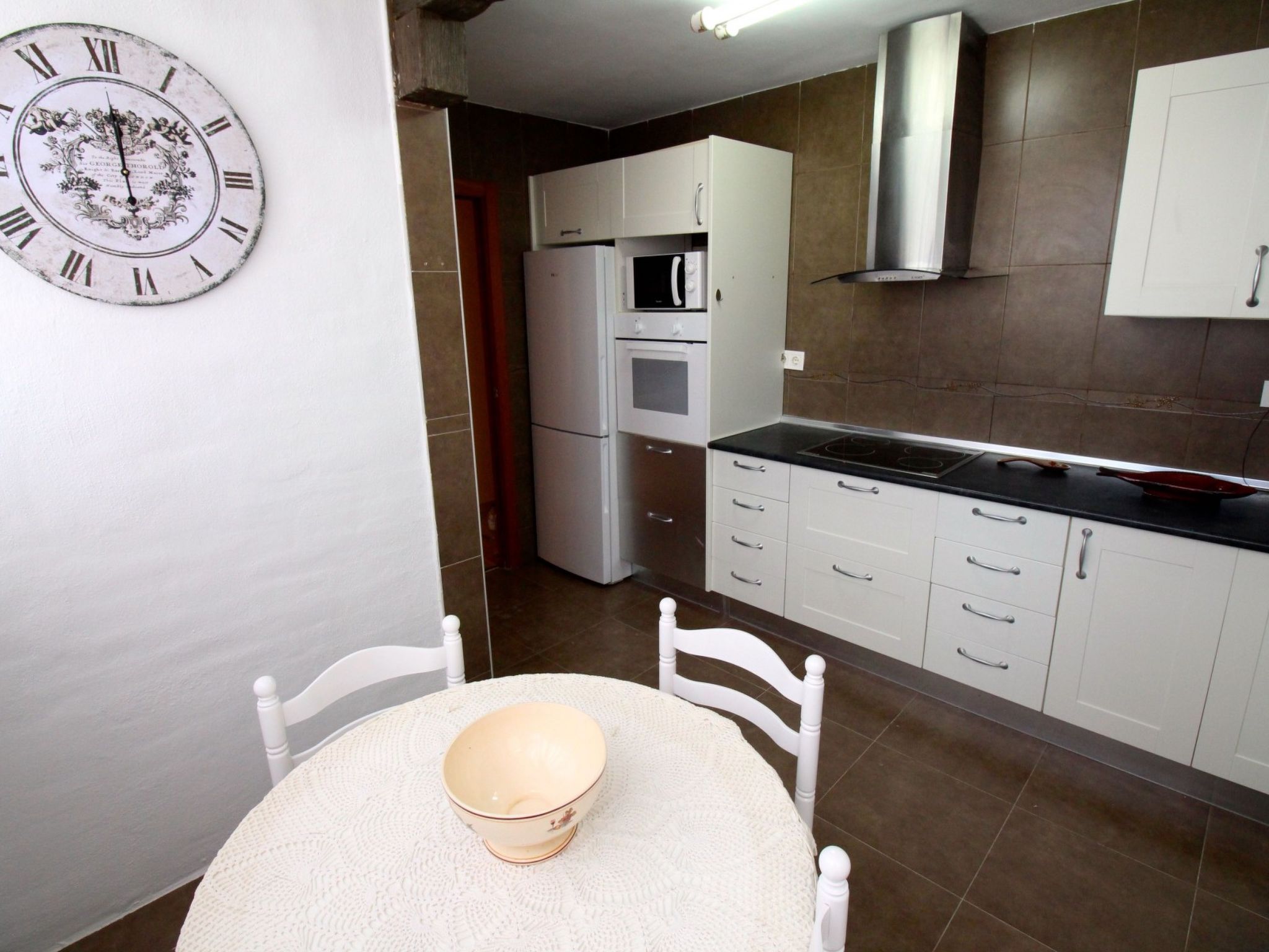 A kitchen with white cabinets, a microwave, oven, and a black countertop. There is a white chair and a table with a bowl. A wall clock is visible.