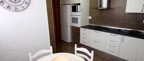 A kitchen with white cabinets, a microwave, oven, and a black countertop. There is a white chair and a table with a bowl. A wall clock is visible.