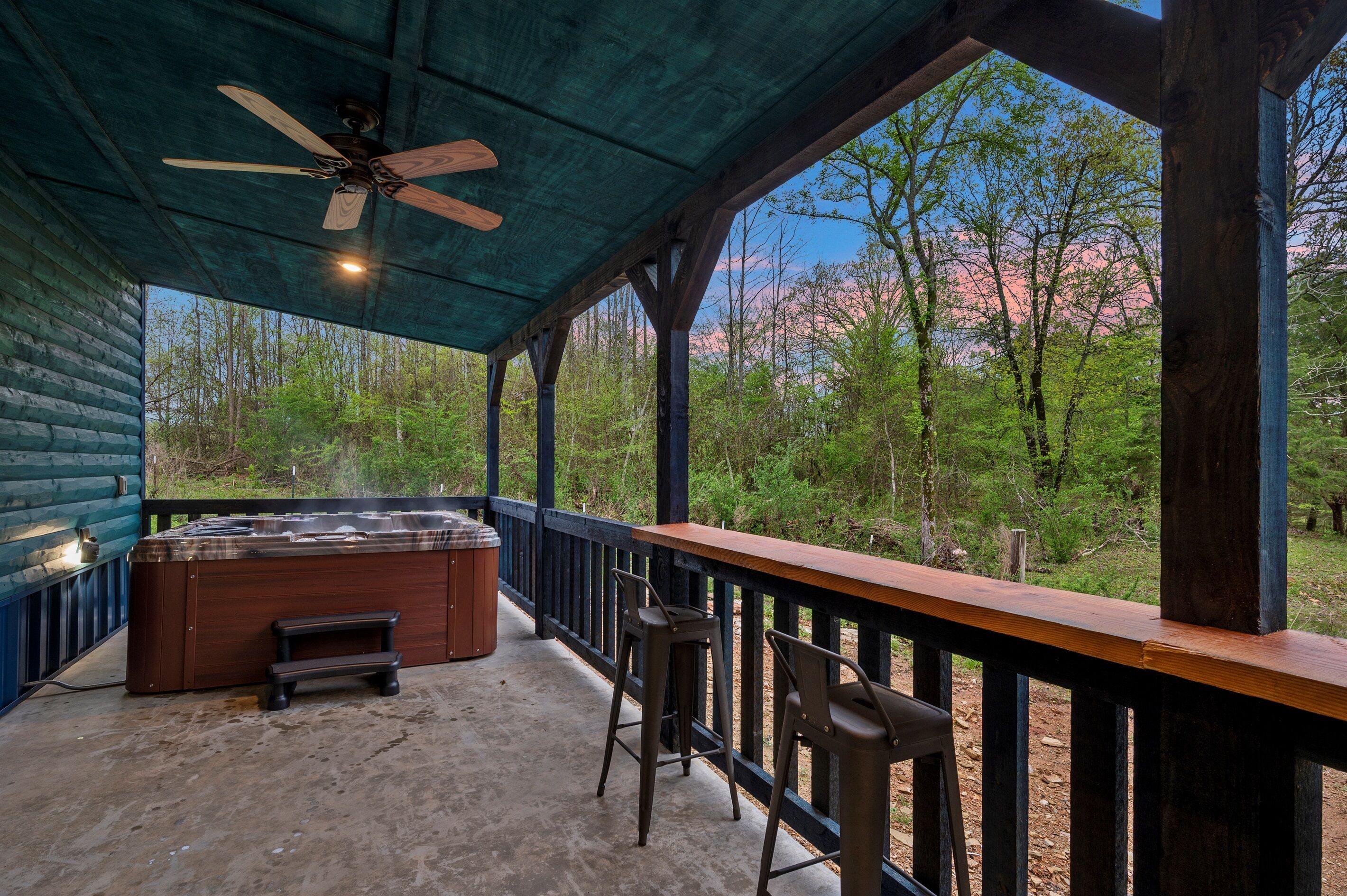 A covered outdoor area with a hot tub, wooden deck, and a view of a forest.