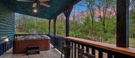 A covered outdoor area with a hot tub, wooden deck, and a view of a forest.