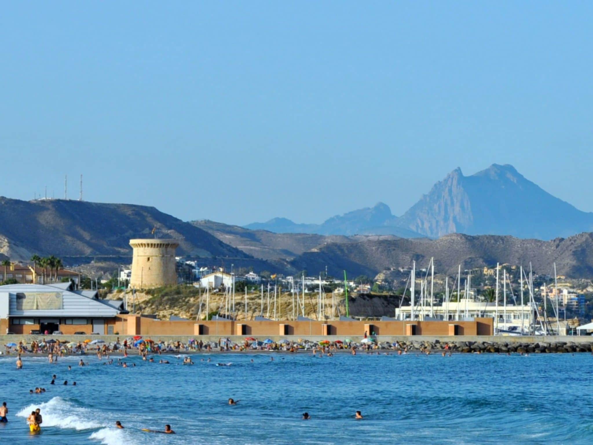 A coastal scene with a tower, a marina, and mountains in the background.