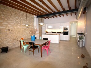 Un comedor con pared de piedra, vigas de madera, suelo de baldosas y una cocina al fondo.
