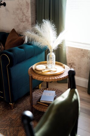 Ein smaragdgrünes Sofa, ein Korbstuhl mit einer Vase und Kerzen, ein Sessel und ein Buch auf einem gemusterten Teppich.
