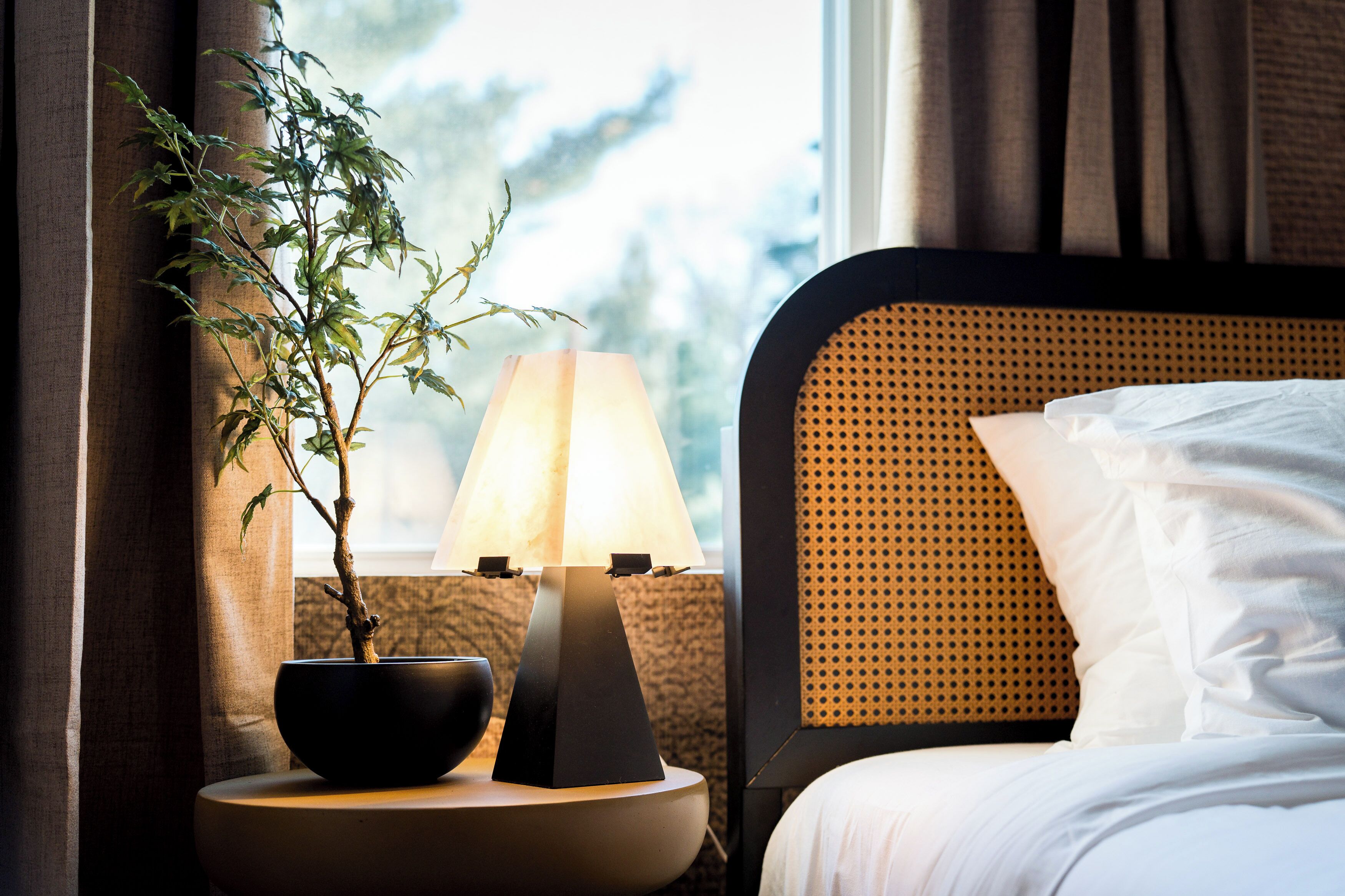 A hotel room with a bed, a bedside table with a lamp, a potted plant, and a window with curtains.