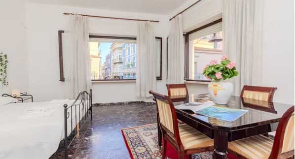 Ein Zimmer mit einem Bett, einem Esstisch mit Stühlen, einer Vase mit Blumen und Blick auf Gebäude durch ein Fenster.