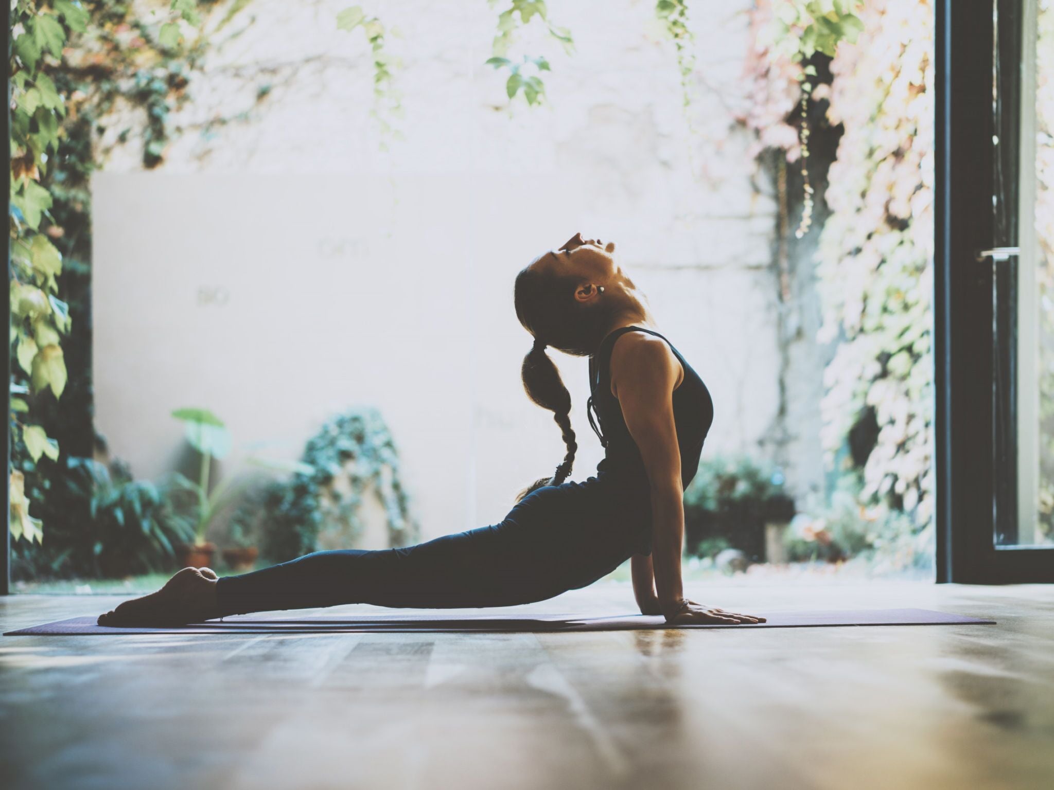 Eine Person führt eine Yoga-Pose auf einer Matte in einem Zimmer mit großem Fenster und Pflanzen aus.