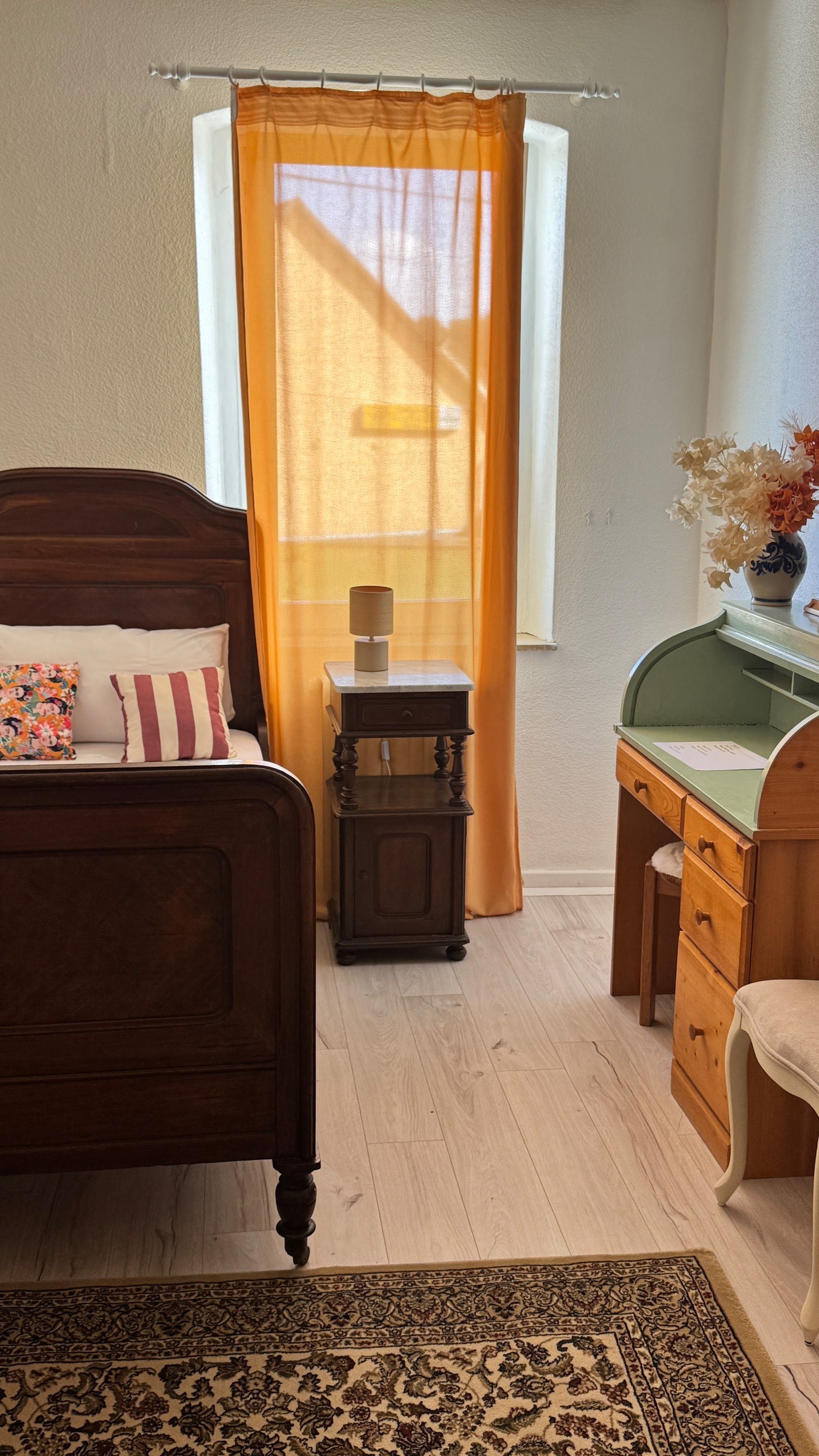 Un dormitorio con cama, mesita de noche, escritorio, silla y ventana con cortinas.