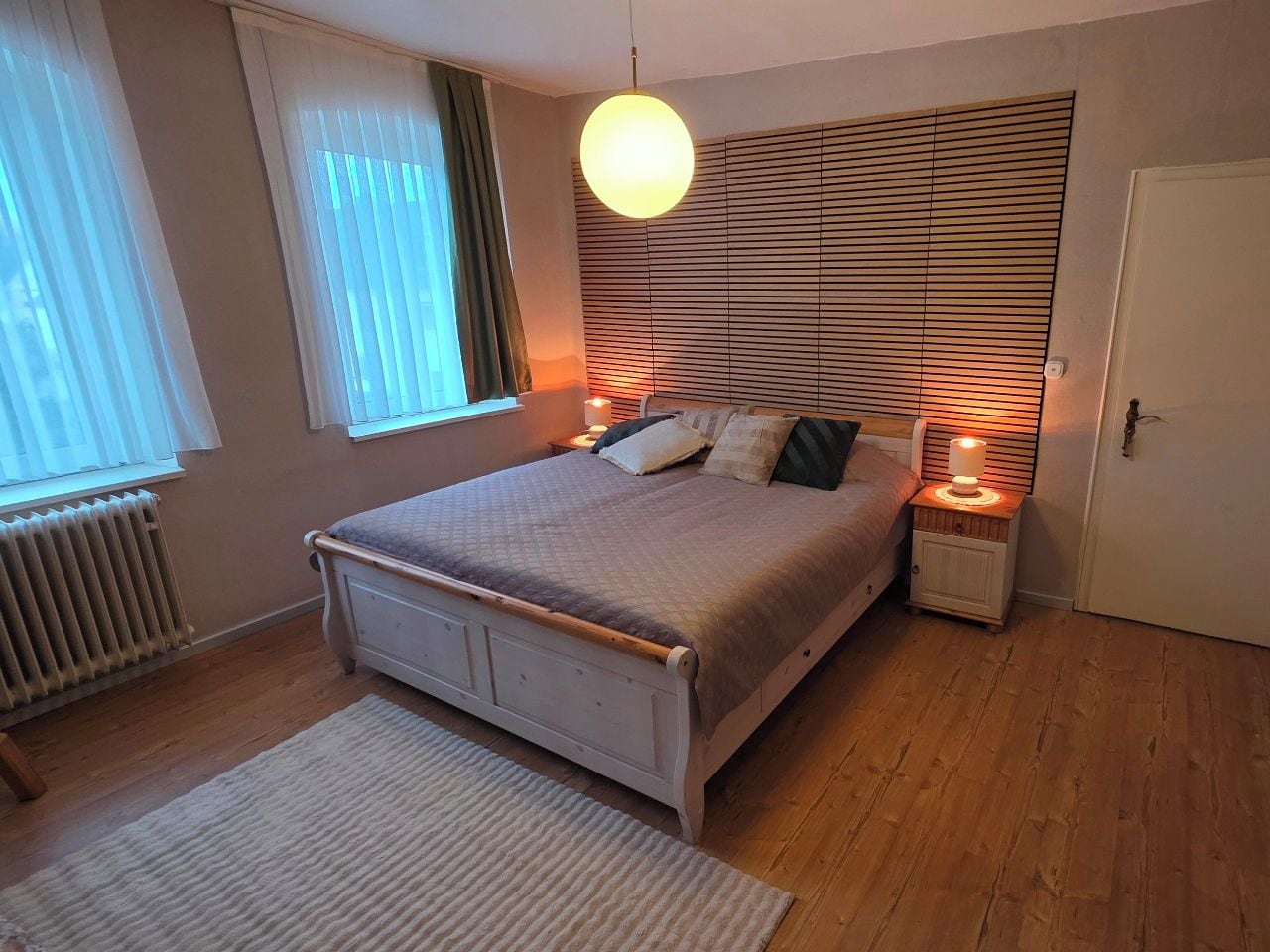 Een slaapkamer met een bed, nachtlampjes, een radiator en een raam met gordijnen.