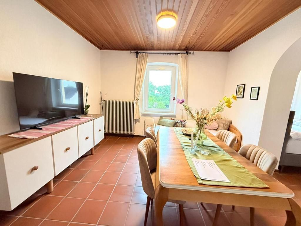 Een eethoek met een houten tafel, stoelen, een televisie op een kast, een raam met gordijnen en een radiator.