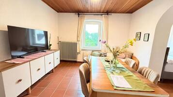 Een eethoek met een houten tafel, stoelen, een televisie op een kast, een raam met gordijnen en een radiator.
