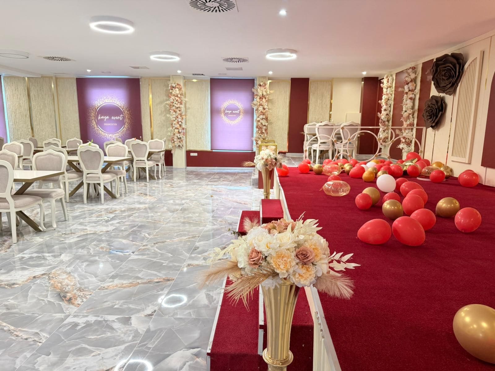 Un salón de banquetes con suelo de mármol, sillas blancas, alfombra roja y decoración floral.