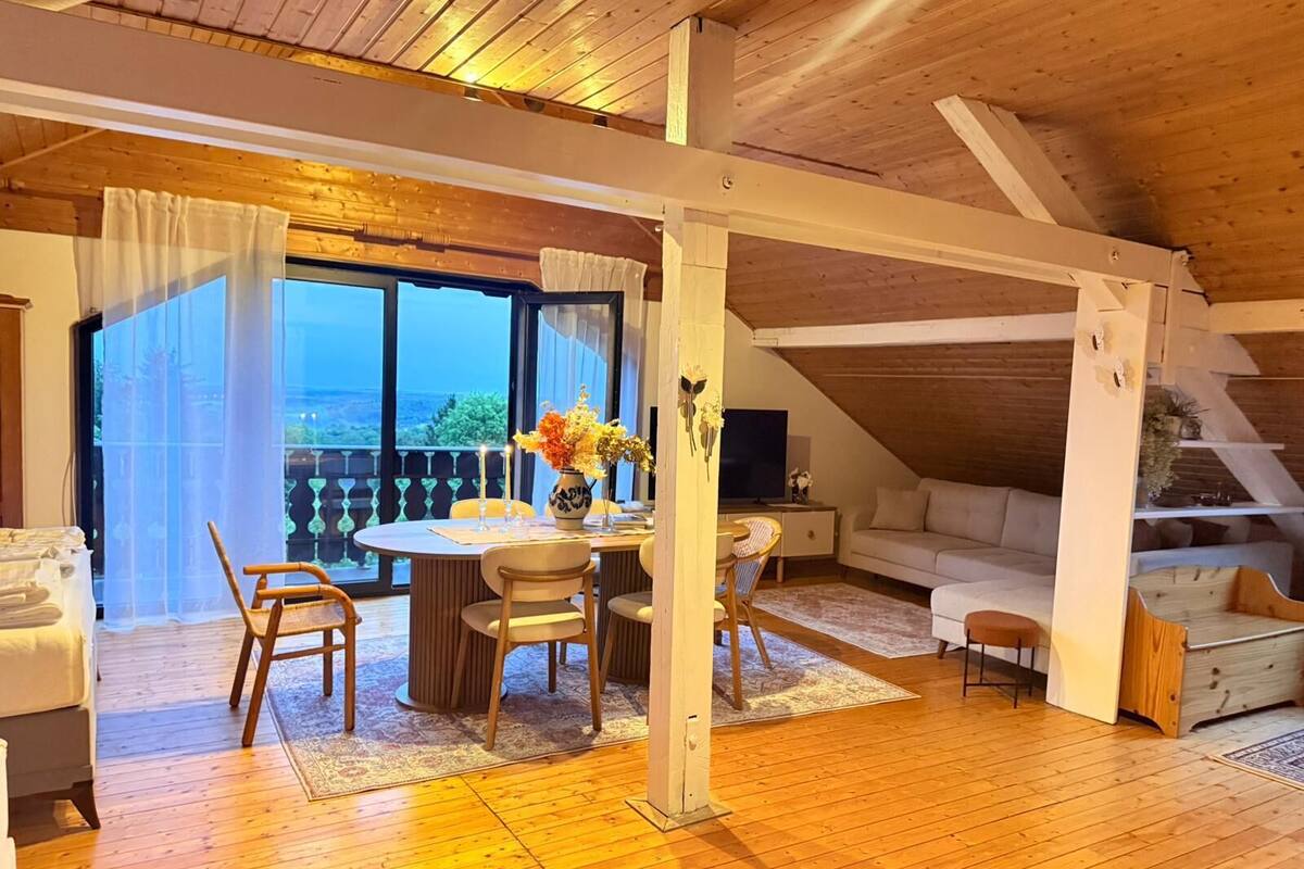A cozy room with a wooden ceiling, a dining area with a table and chairs, a sofa, and a view through large windows.
