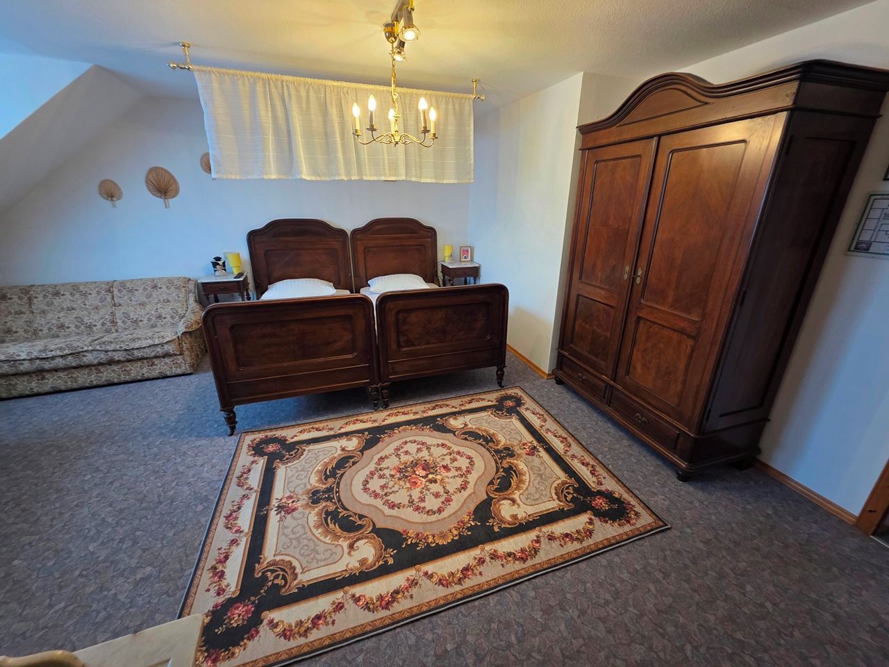 A bedroom with a double bed, a large wooden wardrobe, a chandelier, and a decorative rug.