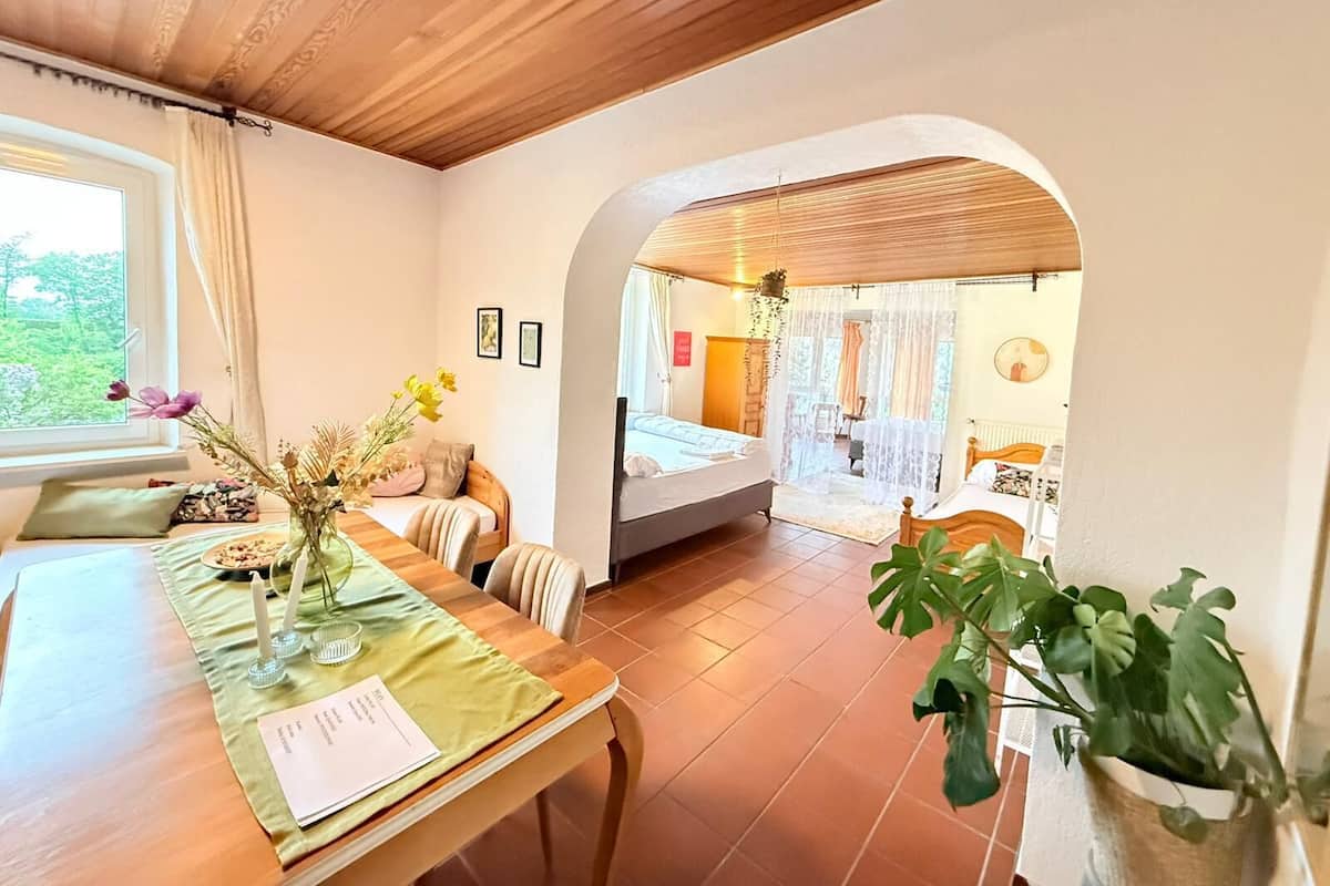 A room with a wooden ceiling, a dining table with a green tablecloth, a bed with a white bedspread, and a large plant.