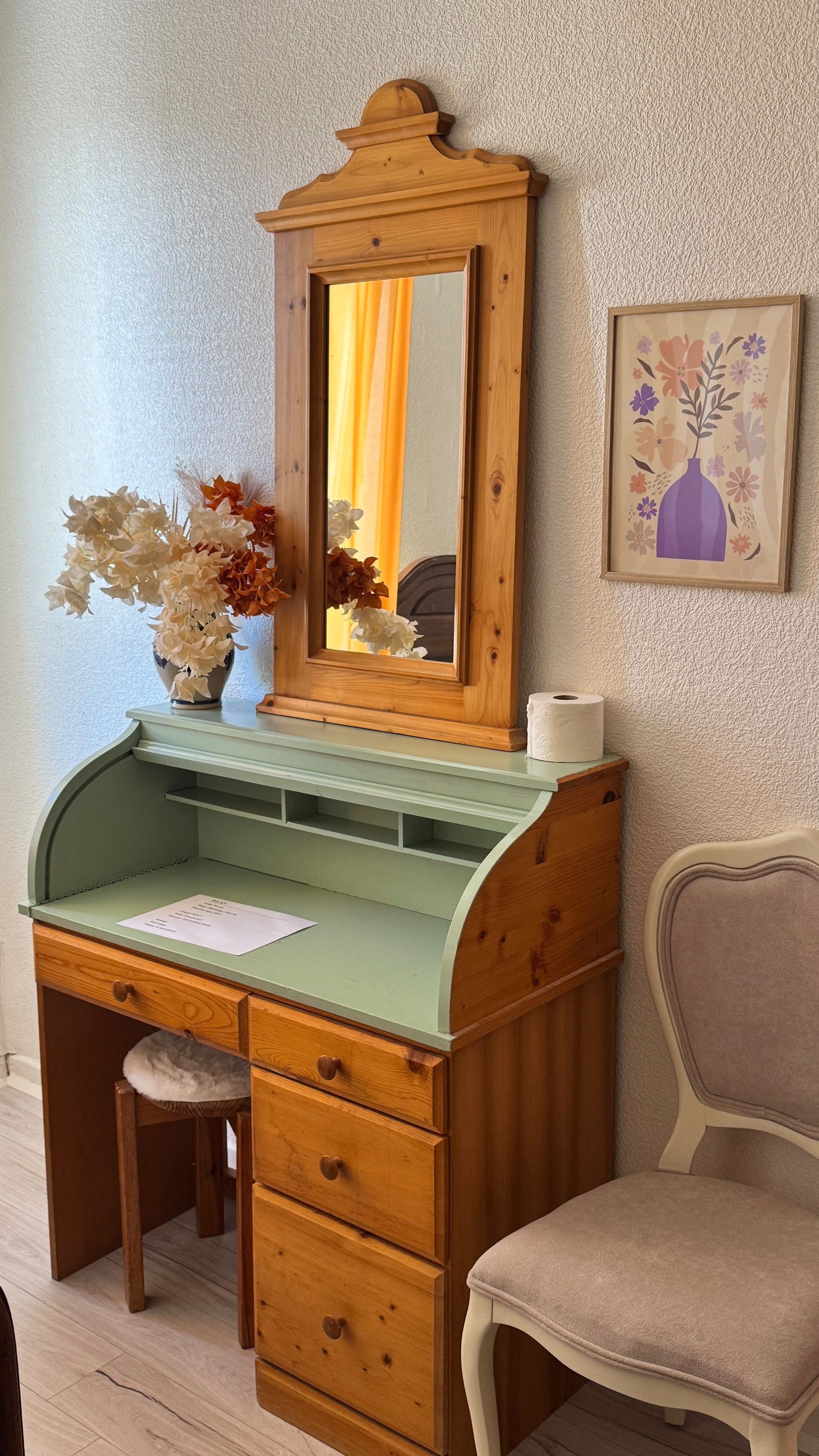 A wooden desk with a mirror, a vase with flowers, and a chair.