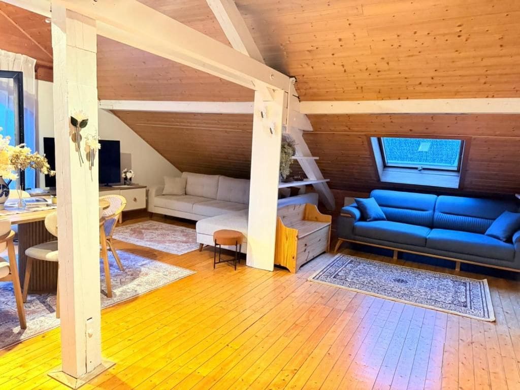 Ein gemütliches Dachzimmer mit Holzbalkendecke, einem weißen Sofa, einem blauen Sofa, einem Esstisch mit Stühlen und einem kleinen Fenster.