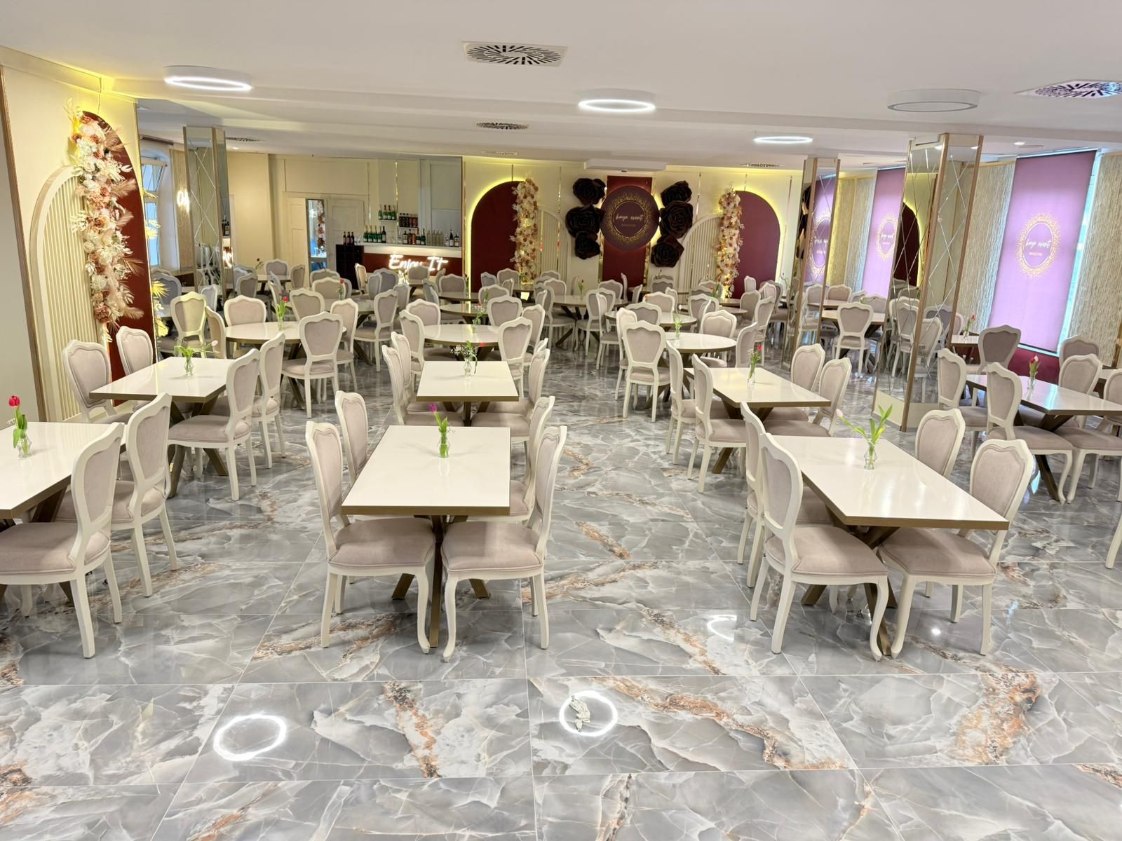 Un vaste espace dédié à la salle à manger, avec un sol en marbre, des tables et des chaises blanches, et un bar en arrière-plan.