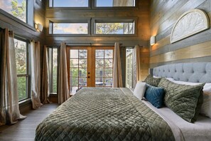 A bedroom with a large bed, a grey upholstered headboard, and a view through a glass door to the outside. - Above the Clouds · New Listing!  Scenic Views, Deck & Game Room (Broken Bow)