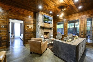A log cabin interior with a stone fireplace, wooden walls, and a television mounted above it. - Above the Clouds · New Listing!  Scenic Views, Deck & Game Room (Broken Bow)