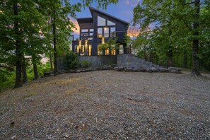 A modern house with a stone foundation, surrounded by a gravel driveway and trees. - Above the Clouds · New Listing!  Scenic Views, Deck & Game Room (Broken Bow)