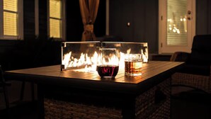 Una mesa con una copa de vino tinto y un vaso de líquido ámbar, una chimenea con llamas, y una silla de mimbre en una habitación con poca luz.